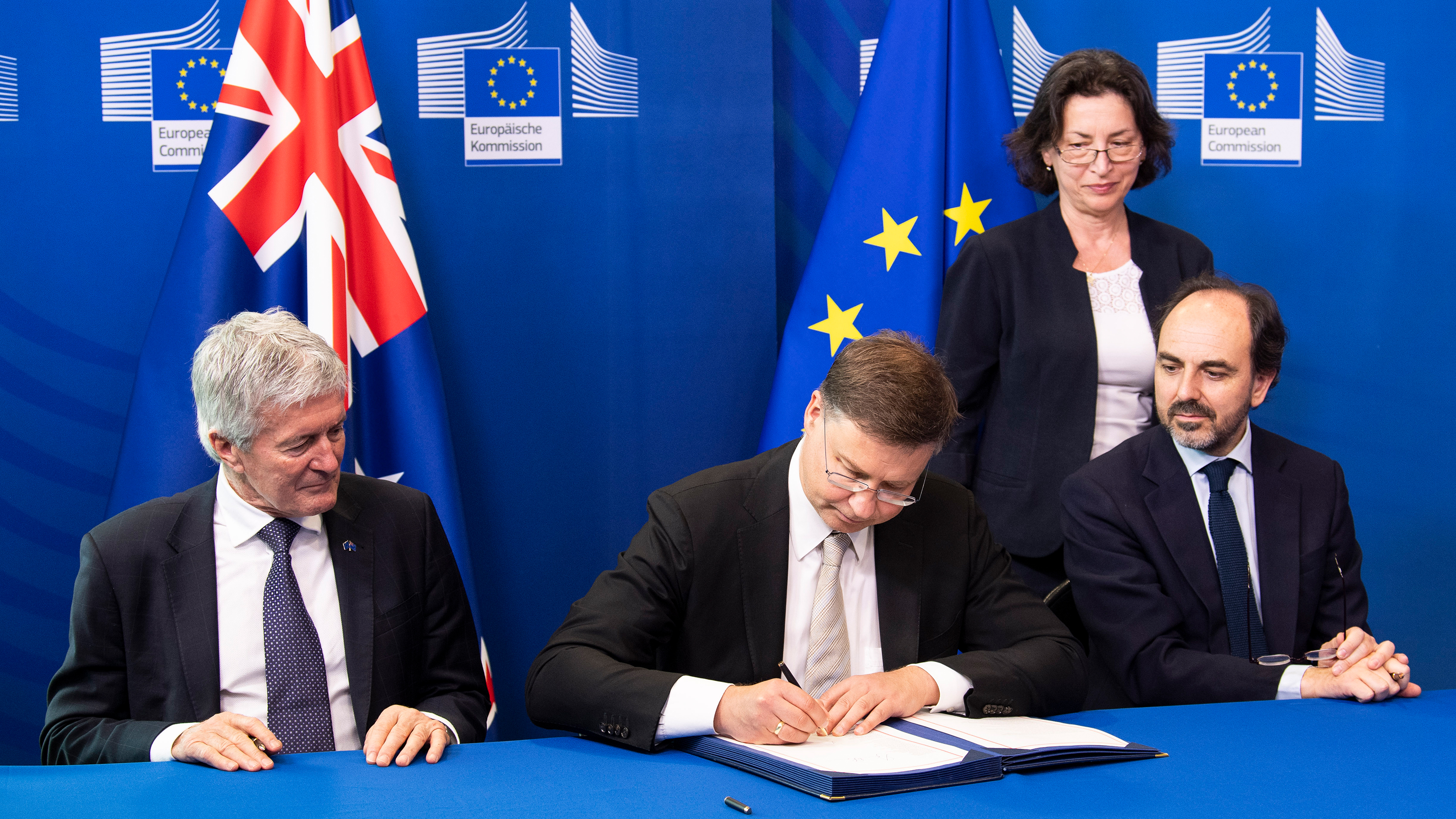 Peron in Anzug sitzt an Tisch und signiert Dokument in Mappe, links und rechts zwei weitere Personen sitzend darauf blickend, im Hintergrund weitere Person stehend vor blauer Wand mit Aufschrift Europäische Kommission