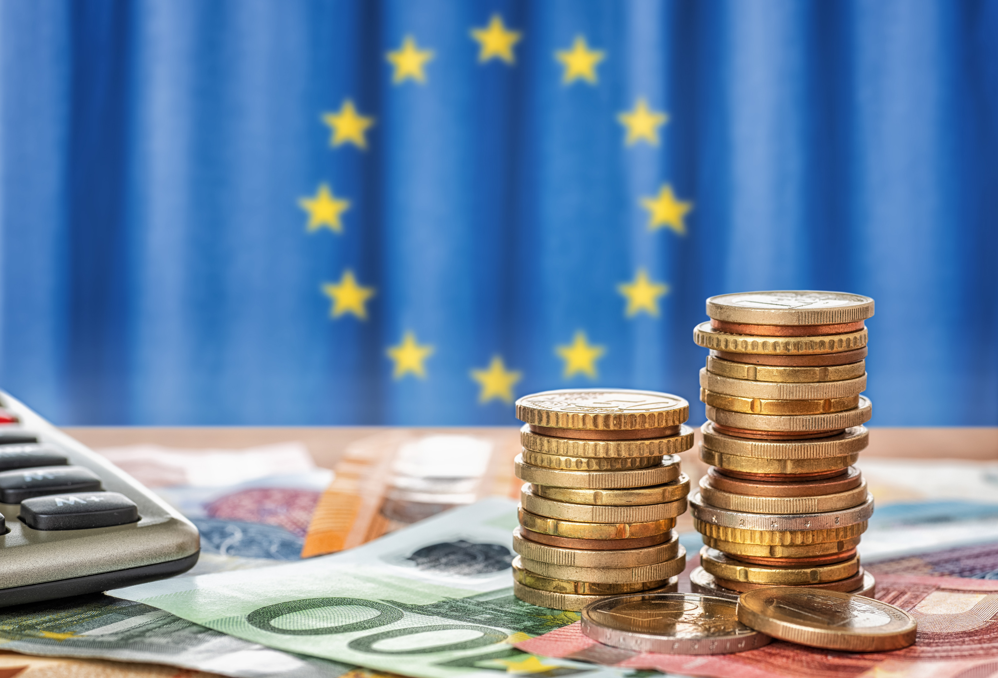 Zu Türmchen gestapelte Euro-Münzen auf Euro-Scheinen platziert, seitlich im Anschnitt ein Taschenrechner, im Hintergrund verschwommen EU-Flagge: blauer Stoff mit gelb in Kreisform verlaufenden Sternen