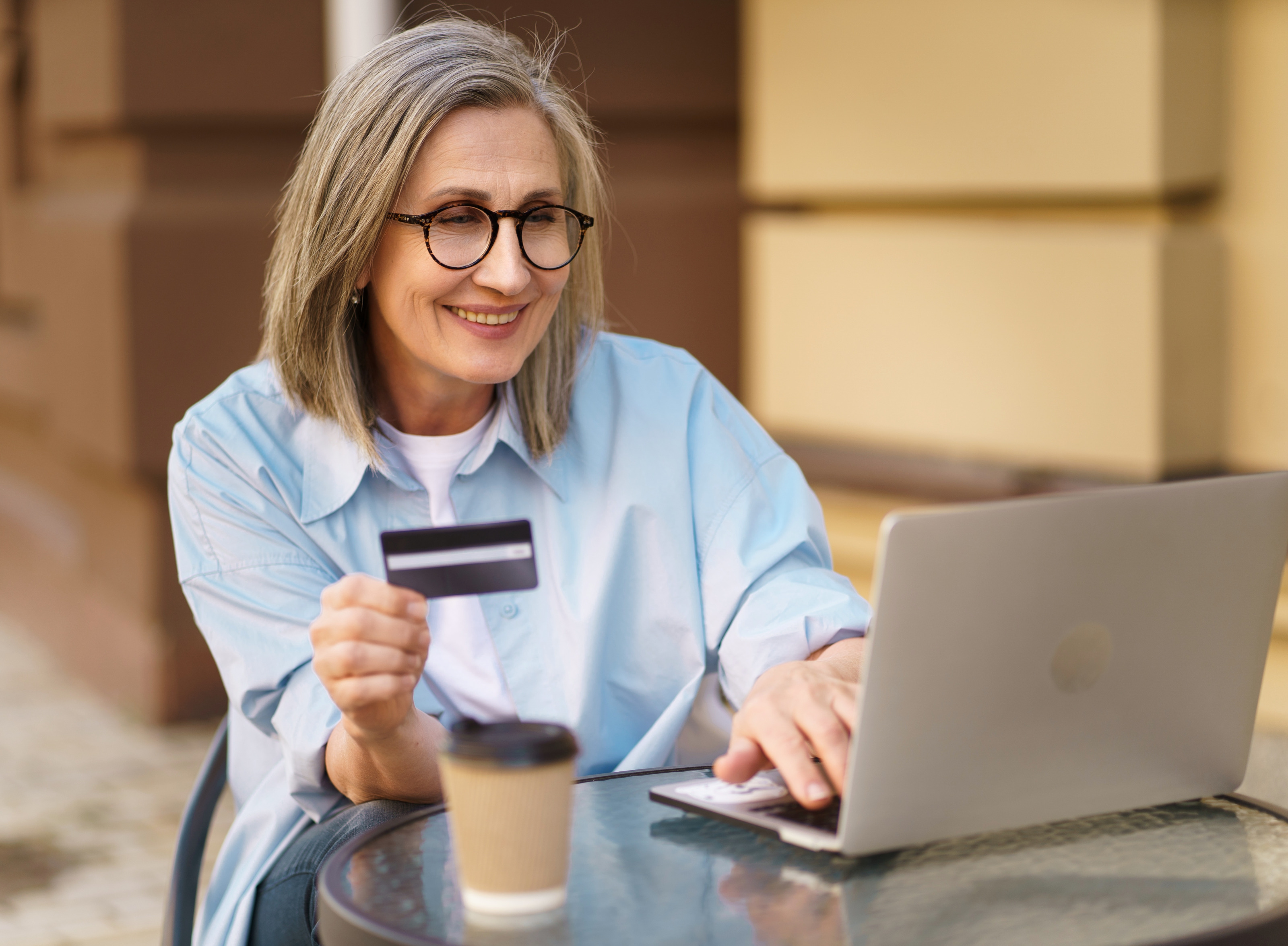 Lächelnde Person mit Brillen Bistrotisch vor aufgeklappten Laptop sitzend hält Bankkarte in Händen und blickt auf Monitor, auf Tisch verschwommen im Vordergrund Kaffeebecher
