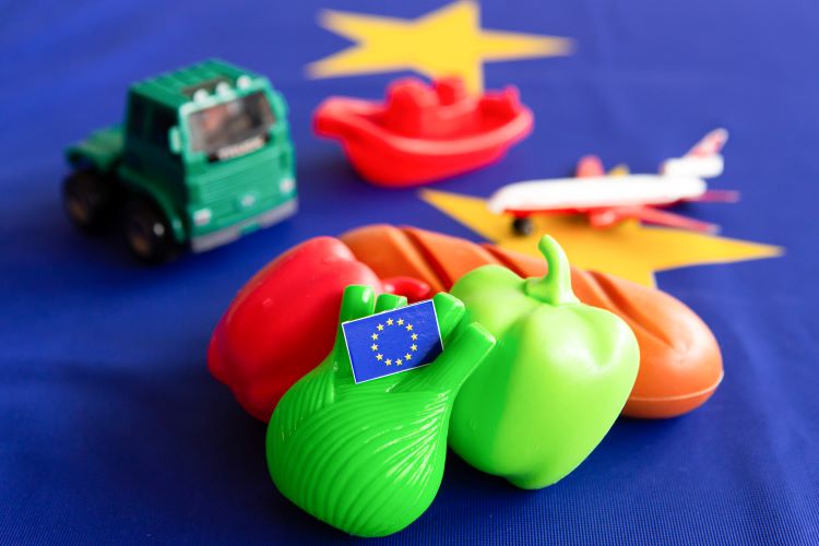 Kleine Plastikmodelle eines Paprikas, eines Baguettes, einer Paprika, eines LKWs, eines Schiffes und eines Flugzeugs liegen auf einer EU-Flagge