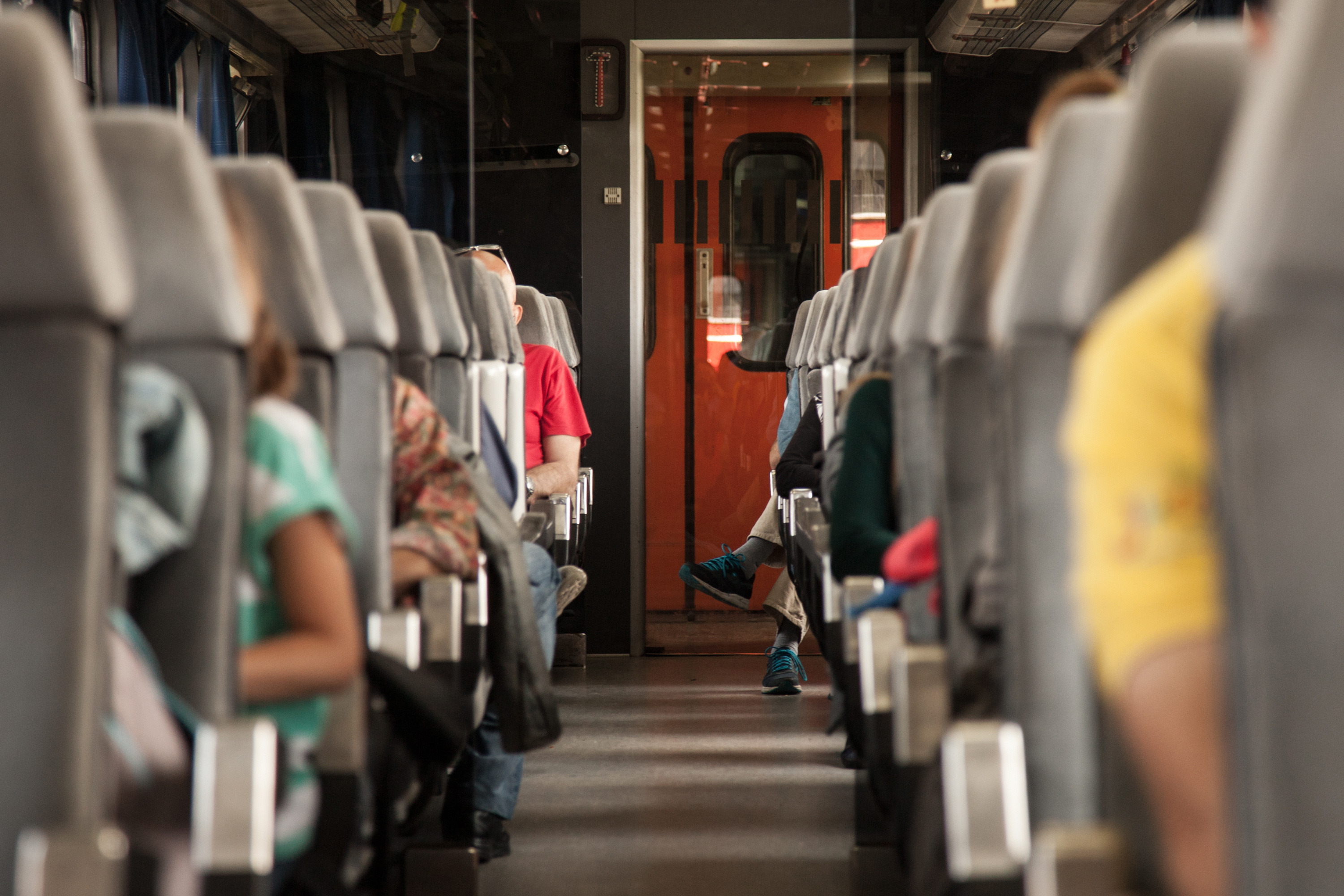 Innenaufnahme eines Zugwagons: Links und Rechts vom Gang graue Sitzplätze in zahlreichen Reihen hintereinander, Menschen im Ausschnitt darauf sitzend erkennbar, im Hintergrund rote Wagontüre